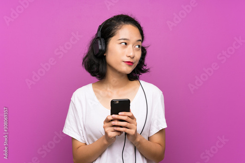 Asian young girl listening music with a mobile over isolated purple wall