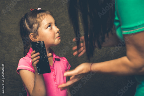 Sad child looks at mom. The concept of strict parenting. Mom punishes her daughter for a broken phone screen. A little girl holds a cracked phone in her hand and looks at her mother plaintively. photo