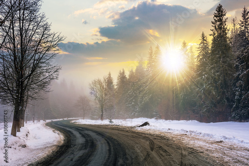 country road through forest at sunset. misty winter weather in evening light. snow on the roadside. cloudy sky