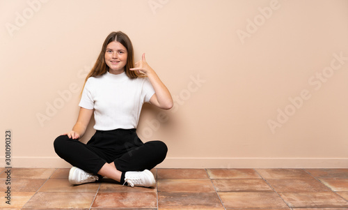 Ukrainian teenager girl sitting on the floor making phone gesture. Call me back sign
