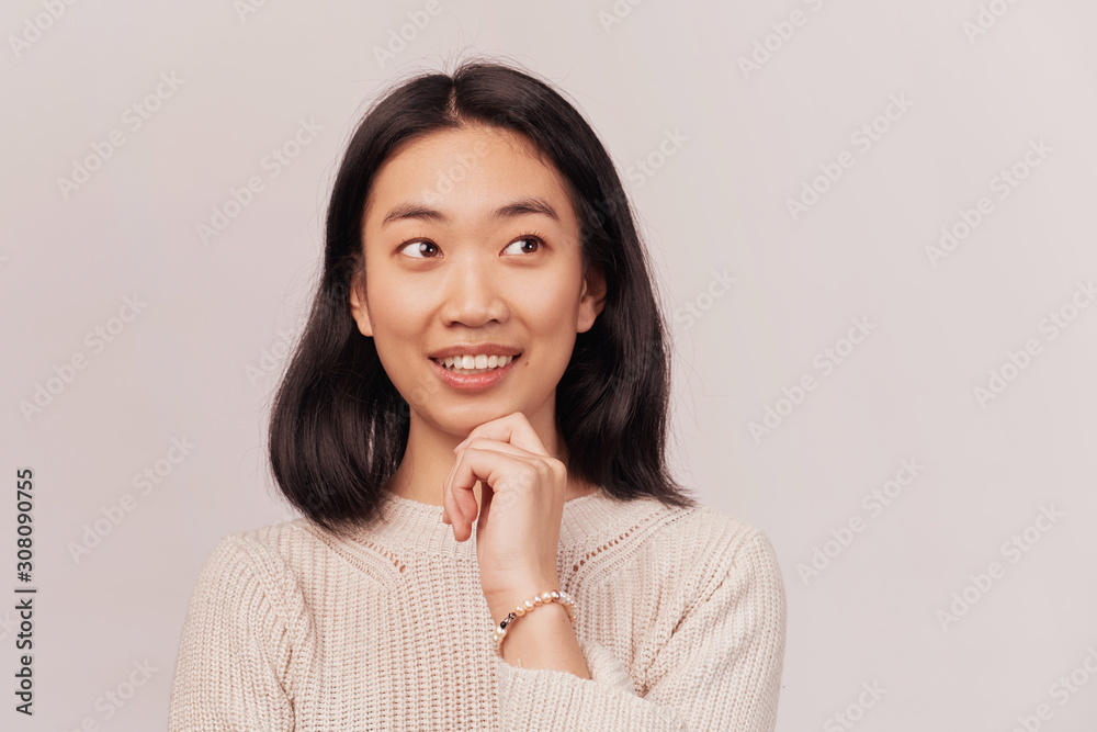 Girl looks up, holding fist on chin, laughs, rejoices. Businesslike young woman Asian appearance with black hair brown eyes dressed in knitted warm sweater stands isolated white background in Studio Girl looks up, holding fist on chin, laughs, rejoices. Businesslike young woman Asian appearance with black hair brown eyes dressed in knitted warm sweater stands isolated white background in Studio