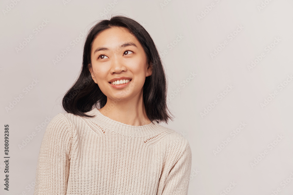 Portrait of charming smiling young girl looking up aside to place of advertising. Businesslike young woman Asian appearance dressed in knitted warm sweater stands isolated white background in Studio. Portrait of charming smiling young girl looking up aside to place of advertising. Businesslike young woman Asian appearance dressed in knitted warm sweater stands isolated white background in Studio.