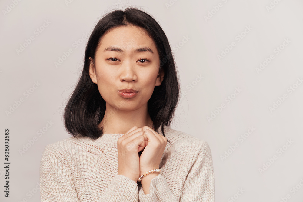 Girl has plaintive expression, frowns, pulls lips. Businesslike young woman Asian appearance with black hair and brown eyes dressed in knitted warm sweater stands isolated white background in Studio. Girl has plaintive expression, frowns, pulls lips. Businesslike young woman Asian appearance with black hair and brown eyes dressed in knitted warm sweater stands isolated white background in Studio.