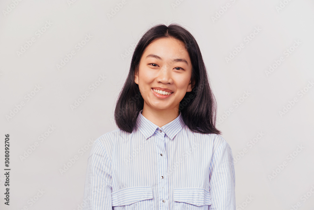 Happy joyful woman laughs and puts hand to chin. Businesslike beautiful young Asian appearance with black hair and brown eyes dressed in striped office shirt stands isolated white background in Studio Happy joyful woman laughs and puts hand to chin. Businesslike beautiful young Asian appearance with black hair and brown eyes dressed in striped office shirt stands isolated white background in Studio