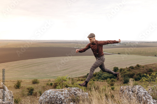 Man in suit and cap jumps on top of rocky hill. The gentleman steps forward  a live moment. Fields and sky on background.