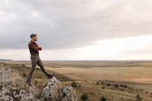 Image of a young farmer who stays on the top of the rock hill and looks into the distance to the beautiful landscape of own fields, thinking about future plans. Optimistic mood