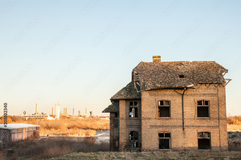 Fototapeta premium Abandoned house facing old factories and warehouses with their distinctive chimneys in Eastern Europe, in Pancevo, Serbia, former Yugoslavia, during a sunny winter afternoon