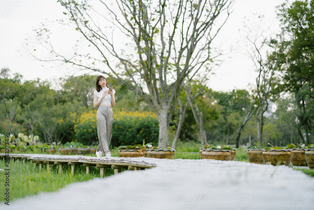 young smiling woman in white clothes with earphones using mobile phone listening to music with her eyes looking at the screen enjoying her moment while strolling on wooden walkway in the park young smiling woman in white clothes with earphones using mobile phone listening to music with her eyes looking at the screen enjoying her moment while strolling on wooden walkway in the park