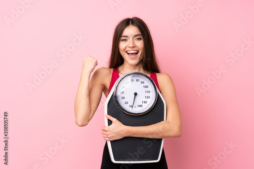 Pretty young girl with weighing machine over isolated pink background with weighing machine and doing victory gesture photo