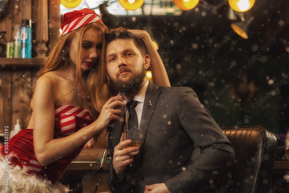 Confident male client in smart suit looking at camera and posing while sexy female stylist shaving him in barbershop. Young woman in santa claus costume working. Concept of new year servicing. Confident male client in smart suit looking at camera and posing while sexy female stylist shaving him in barbershop. Young woman in santa claus costume working. Concept of new year servicing.