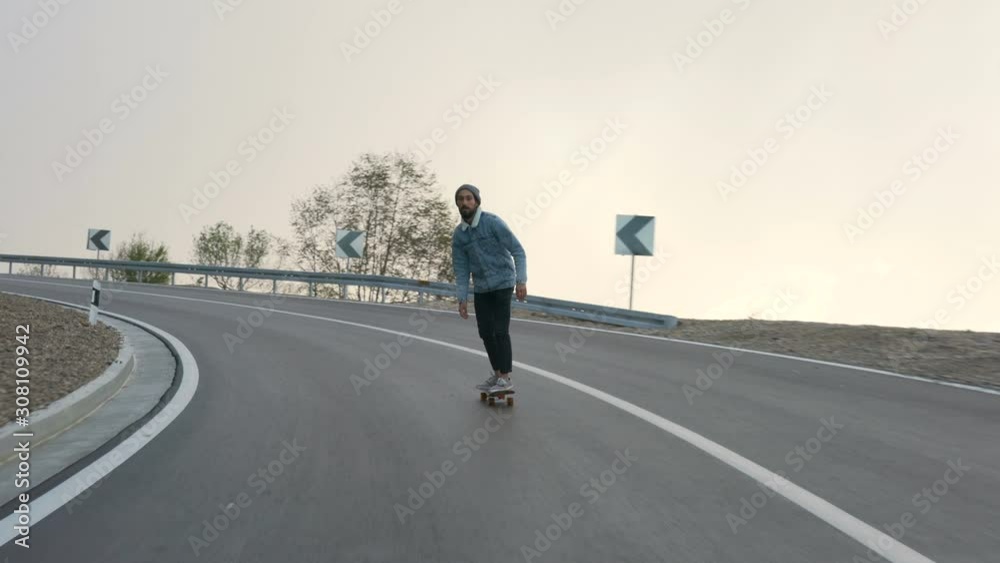young man with a beard riding skateboard cruising downhill on countryside road young man with a beard riding skateboard cruising downhill on countryside road