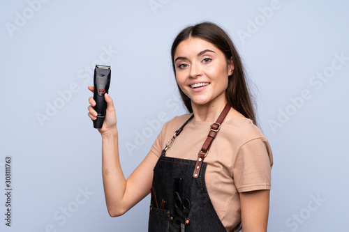 Young woman over isolated blue background with hairdresser or barber dress and holding hair cutting machine