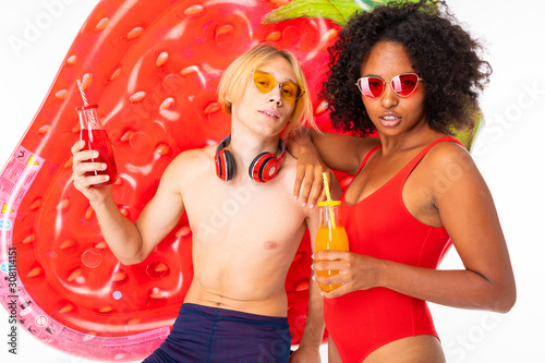 attractive couple european guy and african girl in swimsuits with sunglasses and headphones with cocktails in their hands against the background of swimming mattresses