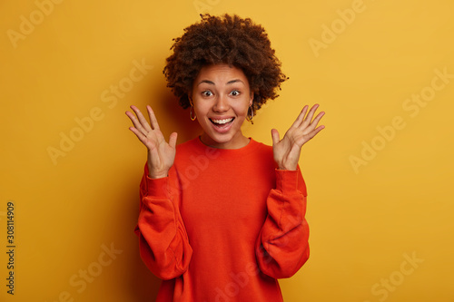 Konstfotografi Half length shot of optimistic curly haired woman keeps palms raised, dressed in red jumper, feels excited by hearing excellent news, smiles broadly, stands against yellow background