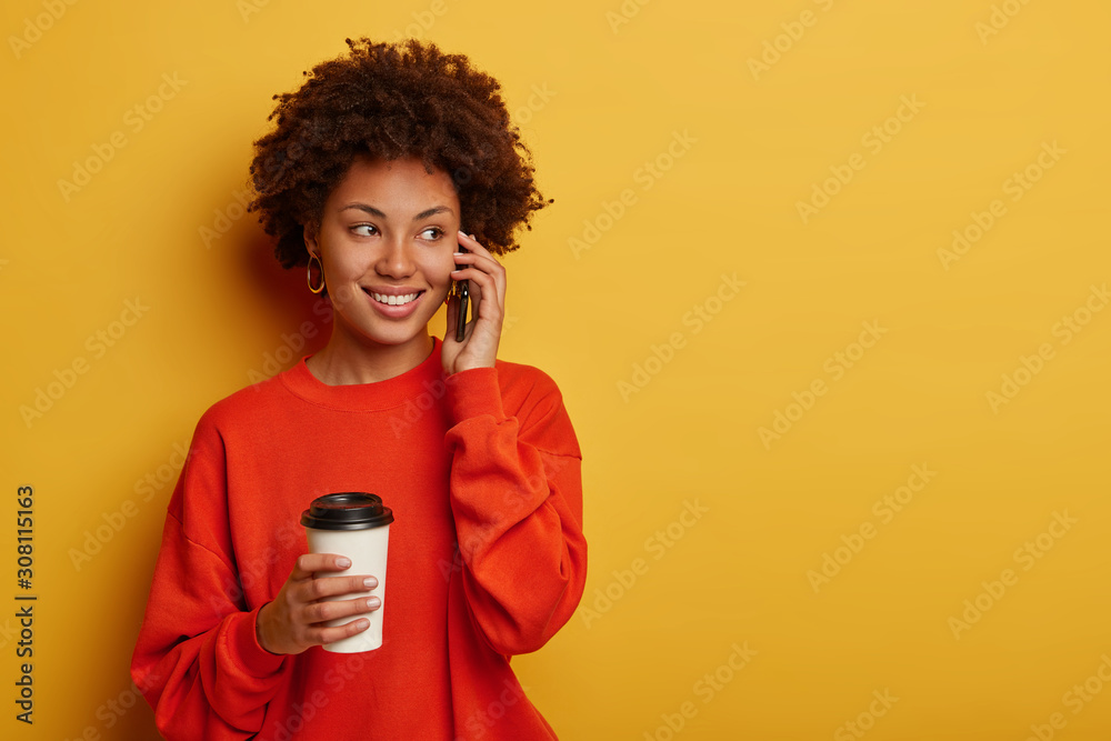 Enthusiastic young Afro girl has phone conversation, speaks with friend and hears funny joke, dressed in casual clothing, drinks aromatic beverage from paper cup looks aside models against yellow wall Enthusiastic young Afro girl has phone conversation, speaks with friend and hears funny joke, dressed in casual clothing, drinks aromatic beverage from paper cup looks aside models against yellow wall