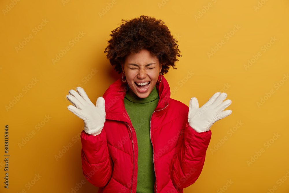 Beautiful dark skinned woman with curly hair, wears winter outerwear, white gloves, expresses happiness, exclaims from pleasure, isolated over yellow studio wall. People and emotions concept Beautiful dark skinned woman with curly hair, wears winter outerwear, white gloves, expresses happiness, exclaims from pleasure, isolated over yellow studio wall. People and emotions concept