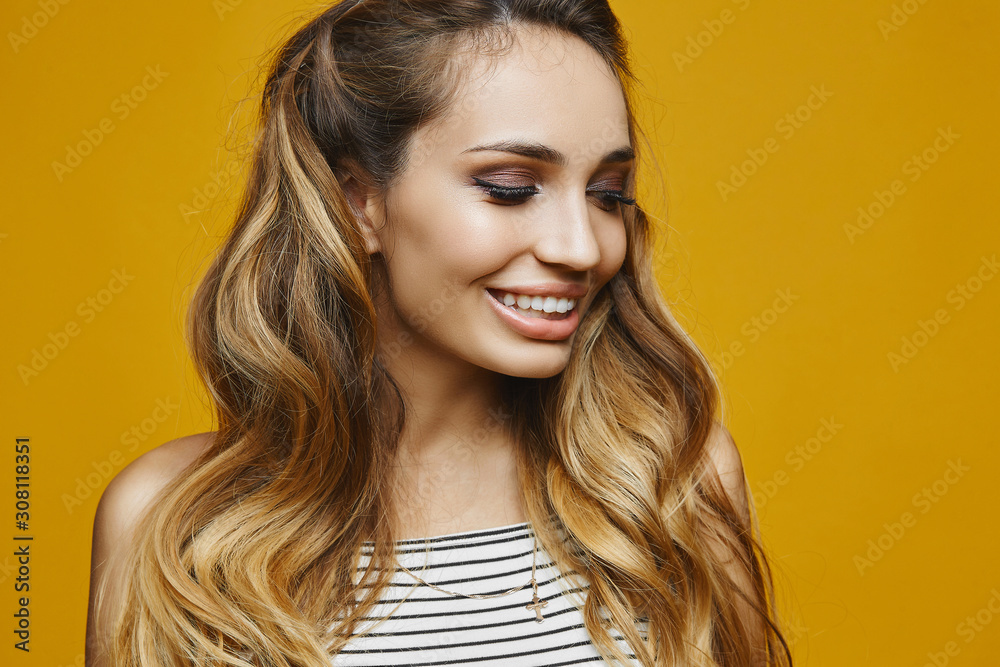 Naklejka premium A fashionable young woman with blond hair and perfect trendy makeup in a striped dress isolated at the yellow background. Beautiful blonde model girl with full lips and a wide smile. Fashion blogger