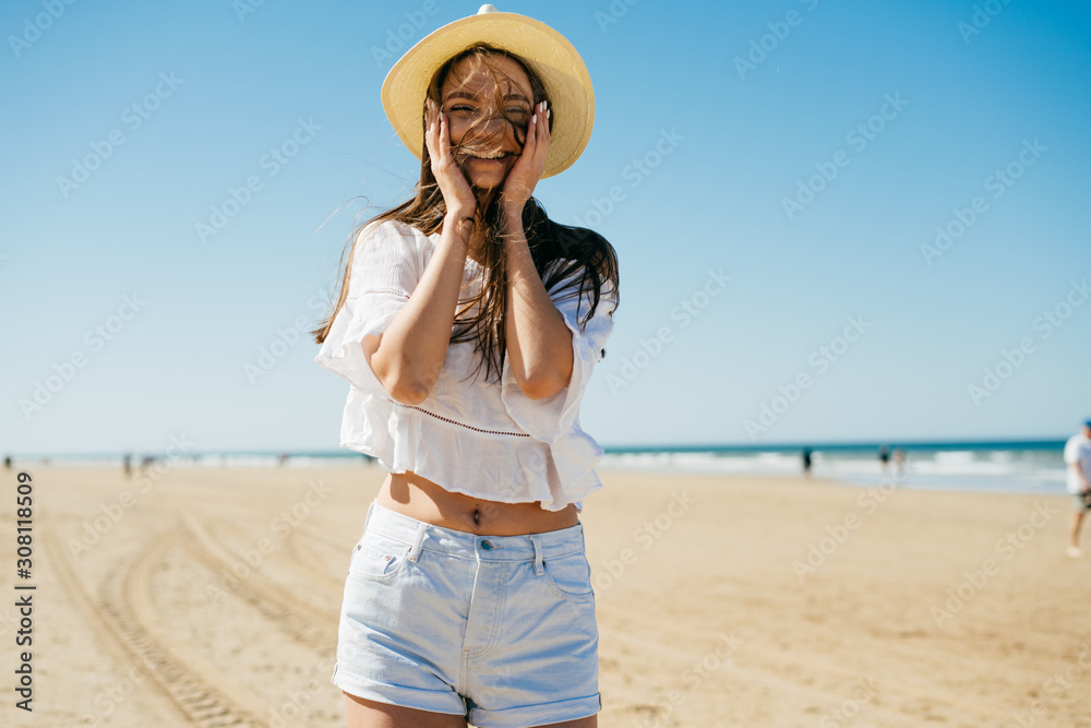 girl laughs and joyfully put her hands to her cheeks, stands on a sandy beach