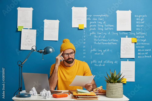 Fototapet Serious male employee or freelancer considers paper document, wears yellow hat a