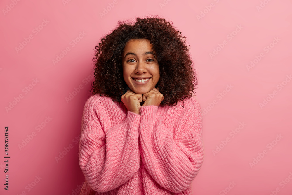 Joyful dark skinned model holds hands under chin, stands casually against pink background, wears oversized jumper, awaits for something pleasant, has beaming smile. People and emotions concept Joyful dark skinned model holds hands under chin, stands casually against pink background, wears oversized jumper, awaits for something pleasant, has beaming smile. People and emotions concept
