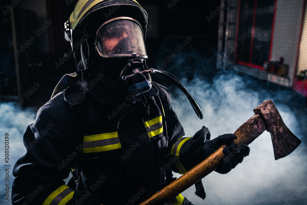 Portrait of a female firefighter while holding an axe and wearing an oxygen mask indoors surrounded by smoke. Portrait of a female firefighter while holding an axe and wearing an oxygen mask indoors surrounded by smoke.