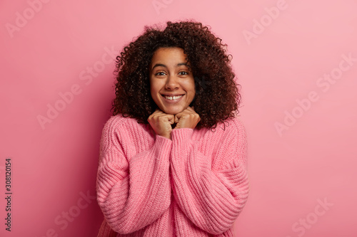 Joyful dark skinned model holds hands under chin, stands casually against pink background, wears oversized jumper, awaits for something pleasant, has beaming smile. People and emotions concept © wayhome.studio 