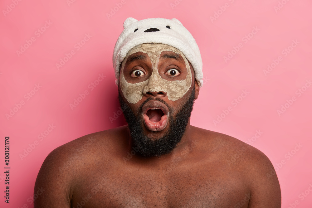 Headshot of stupefied black man applies face mask, stares with eyes popped out, cares about dermatology health, stands bare shoulders, isolated on pink wall. Skin care for men and spa concept Headshot of stupefied black man applies face mask, stares with eyes popped out, cares about dermatology health, stands bare shoulders, isolated on pink wall. Skin care for men and spa concept