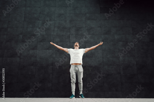 Powerful caucasian handsome fit bearded blond sportsman in tracksuit and in t-shirt holding hands in the air and looking up while standing in front of dark background.