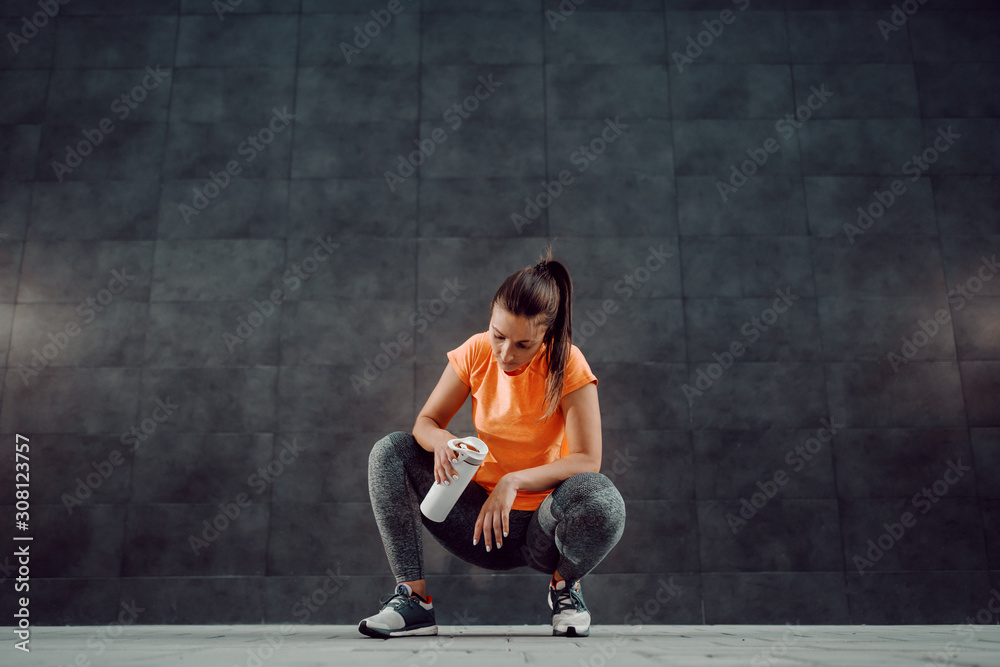 Charming fit caucasian sportswoman in shape dressed in active wear crouching and holding bottle of water. In background is dark wall.