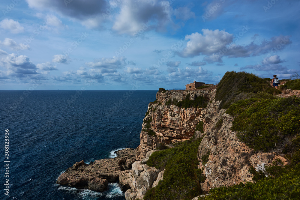 Cliffs and small house by the sea. Hiker walking. Blue sky with clouds. Mallorca, Spain