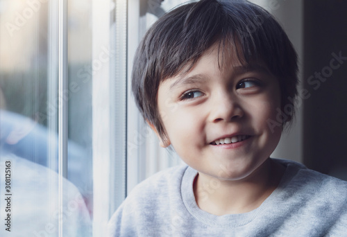 Low key portrait of happy kid looking through window with smiling face,Cute boy playing on his own and making funny face, Child relaxing at home during cold weather outside in Autumn or Winter.