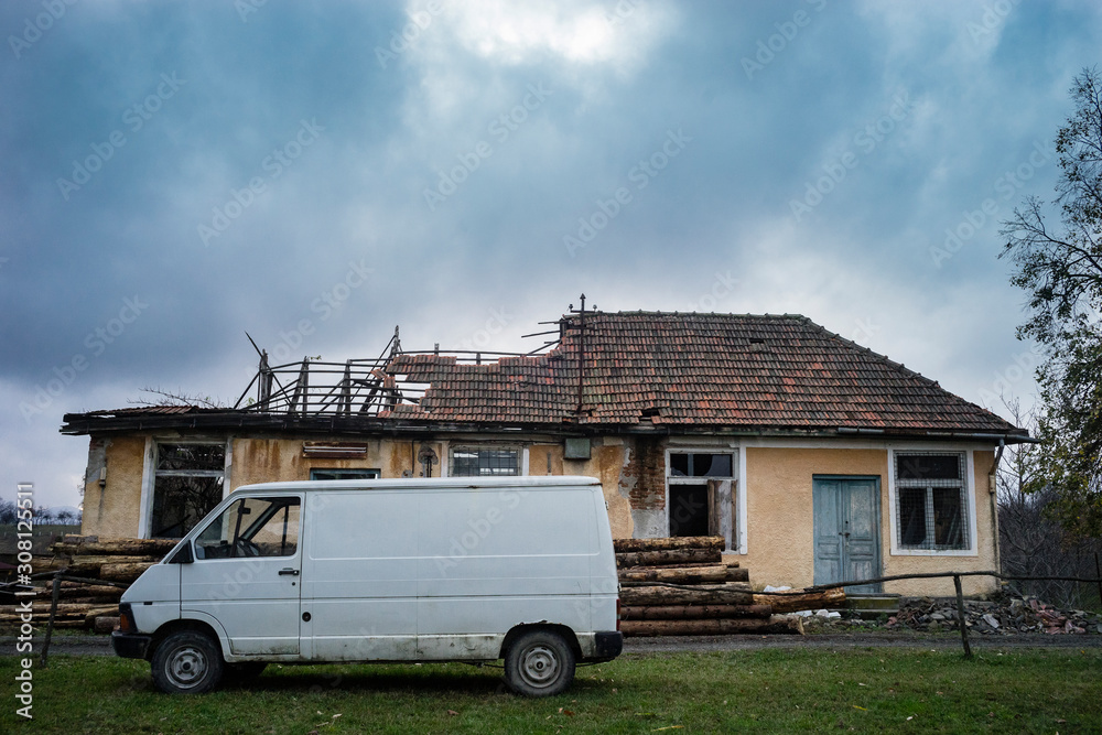 White van parked in front of an abandoned house with part of the roof collapsed.