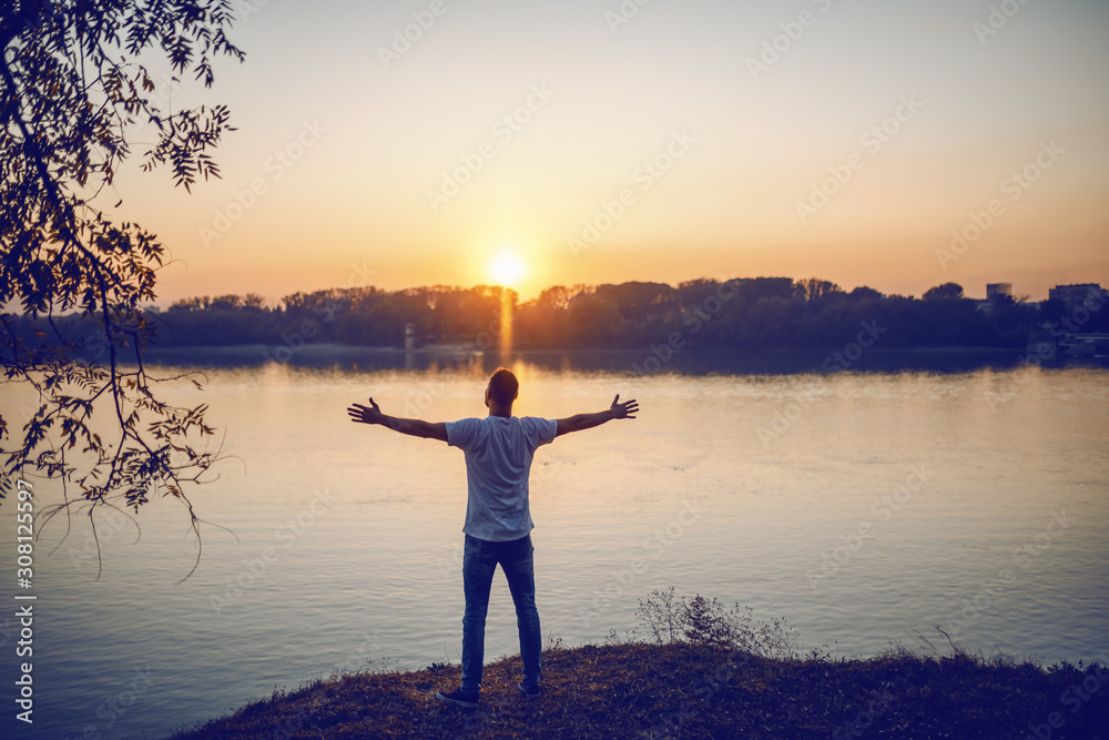 Rear view of handsome caucasian man standing with opened arms on cliff and looking at river and beautiful sunset.