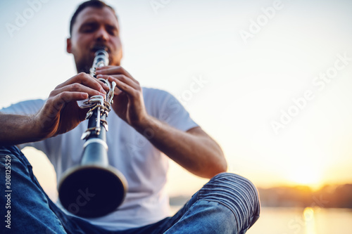 Foto Young handsome bearded caucasian blond man sitting on cliff and playing clarinet