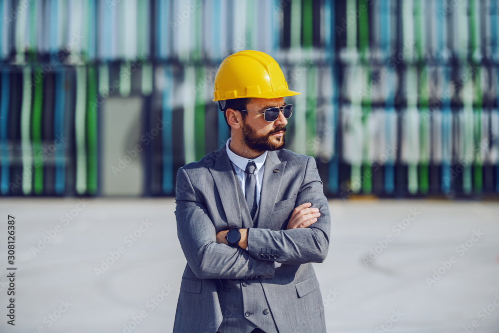 Elegant serious Caucasian bearded businessman in suit, with sunglasses and helmet on head standing on construction site with arms crossed and supervising the works. Elegant serious Caucasian bearded businessman in suit, with sunglasses and helmet on head standing on construction site with arms crossed and supervising the works.