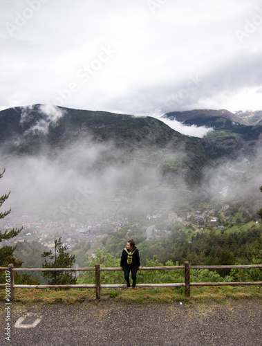 Tourist girl sitting on a wooden fence, with Pyrenees mountains in the background. A small village of Encamp, located in Andorra