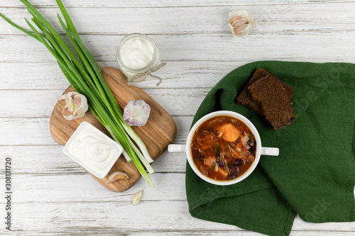 Fototapeta Naklejka Na Ścianę i Meble -  red borsch, a traditional national dish of Ukrainian cuisine. on the stock photo is served in a white pottery with cream, green onions and garlic on a light wooden background. green textile. flat lay