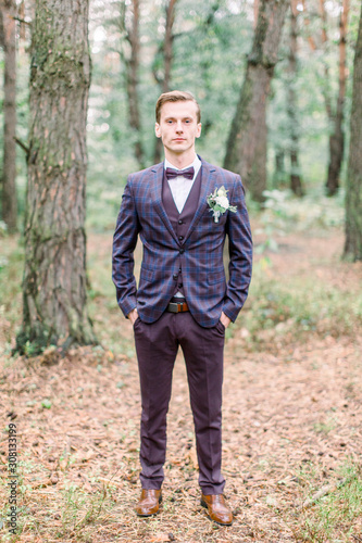 Full length portrait of the groom in a suit and a bow tie. Handsome elegant groom in checkered suit is standing in the forest © sofiko14