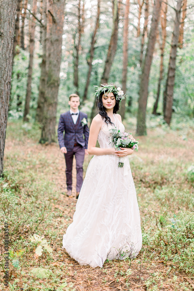 Gorgeous bride in floral wreath holding bouquet, standing in forest. Handsome groom is going to his bride on the background. Rustic wedding outdoors