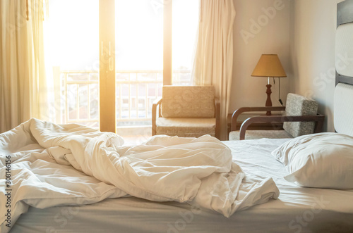 Messy white bedding sheet and white pillow after waking up in the morning with sunshine shining through window with curtains in to the bed room, waking up concept