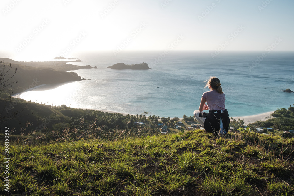 Girl standing on the top of the green mountain, admiring view of a small village with ocean in background on Yasawa island group in Fiji, South Pacific