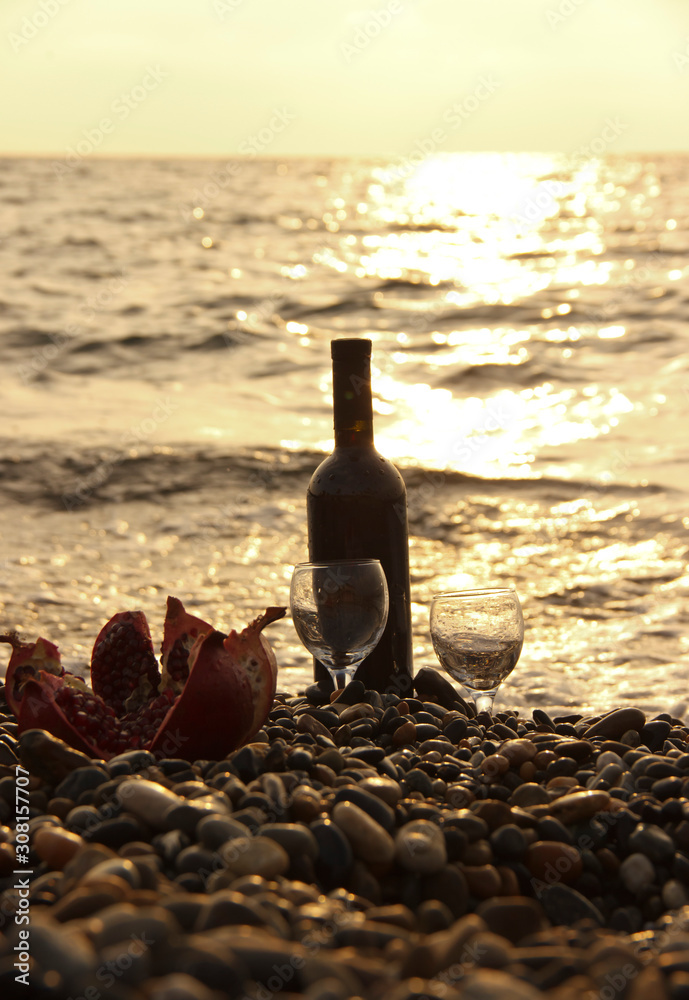 Ripe pomegranate fruit with a glass of wine and a bottle on the beach. Romantic day at the seaside.