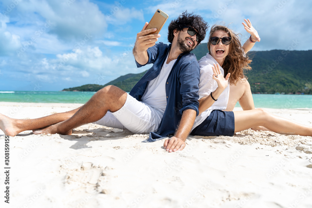 Beautiful young couple joyfully sitting on the white sand by the sea and takes a selfie on a smartphone. Honeymoon Travel and Vacation on the Tropical Ocean Beautiful young couple joyfully sitting on the white sand by the sea and takes a selfie on a smartphone. Honeymoon Travel and Vacation on the Tropical Ocean