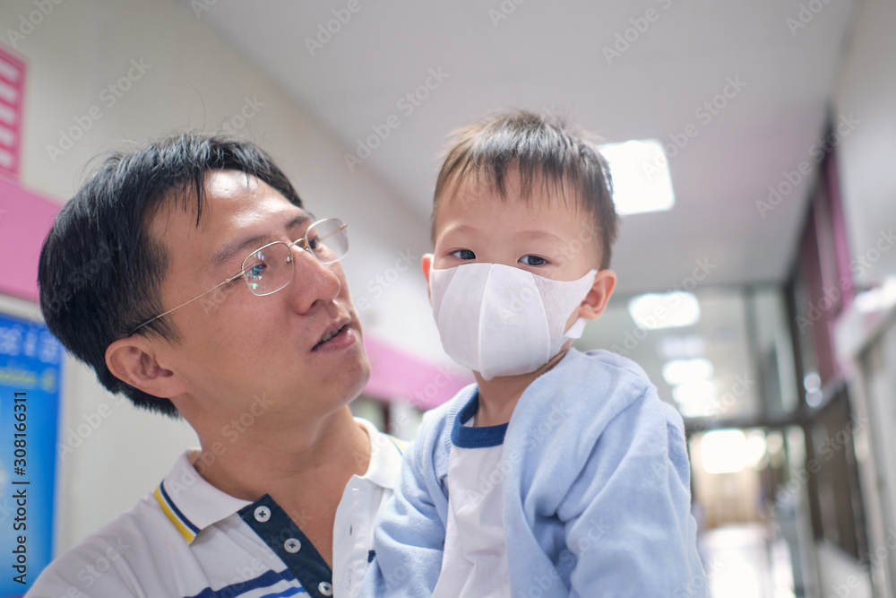 Father holding cute little Asian 3 years old toddler boy child wearing protective medical mask, parent and sick kid, Dad and son waiting to see the doctor at hospital with copy space