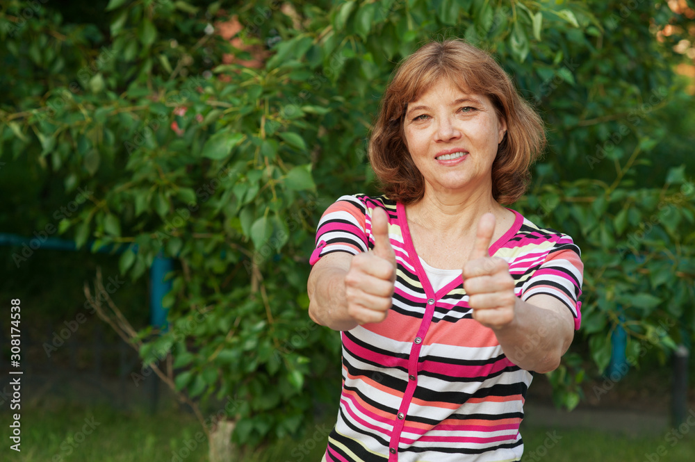 Beautiful happy 60 year old European woman shows two thumbs up on background of green foliage, outdoor copy space. Great and positive mood, healthy lifestyle Beautiful happy 60 year old European woman shows two thumbs up on background of green foliage, outdoor copy space. Great and positive mood, healthy lifestyle
