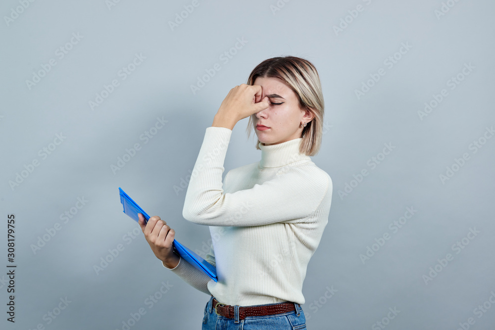 Strict young woman in white shirt, holds folder keeping hand on nose isolated on gray wall background in studio. Healthy fashion lifestyle, people sincere emotions concept. Strict young woman in white shirt, holds folder keeping hand on nose isolated on gray wall background in studio. Healthy fashion lifestyle, people sincere emotions concept.