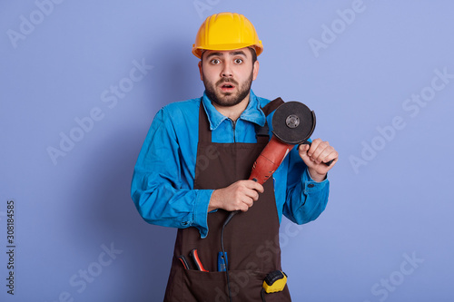 Indoor shot of male builder or manual worker in yellow helmet, holding grinder tool in hands, posing isolated over blue wall background. Repair, construction, building, people and maintenance concept.