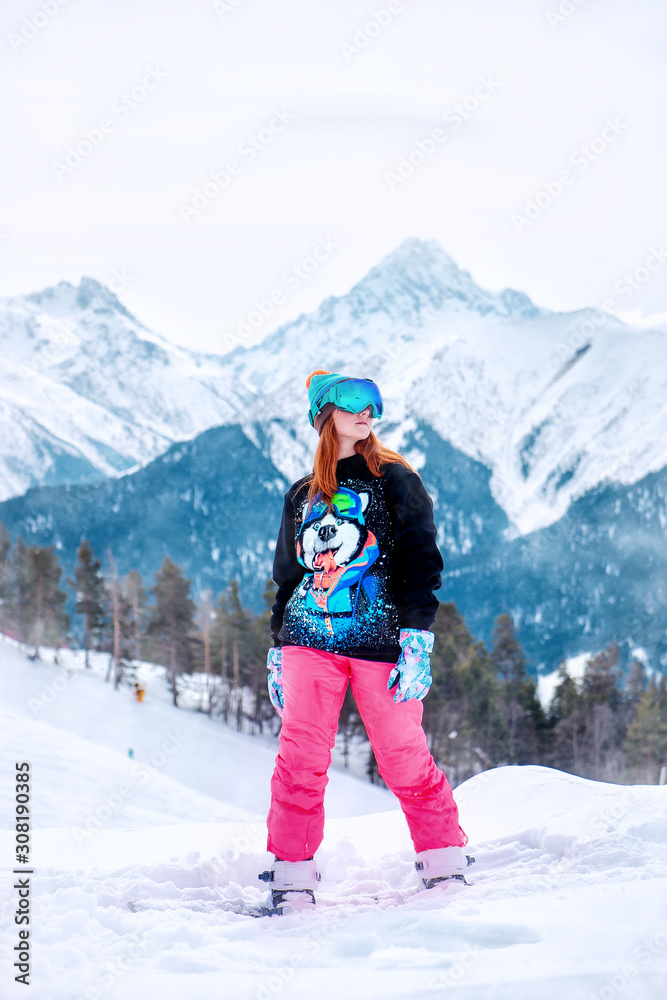 young happy girl stands on a snowboard in bright colored clothes on a background of snowy mountains