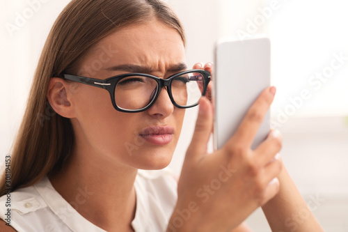 Girl Squinting Eyes Looking At Smartphone Through Eyeglasses Sitting Indoor photo