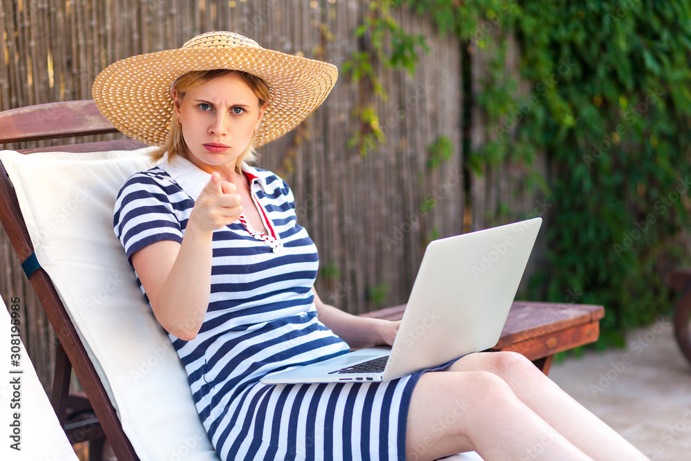 Hey you! Portrait of angry serious young adult freelancer woman in hat and dress sitting on cozy daybed with laptop and pointing finger. Lifestyle concept, outdoor, summer vacation, looking at camera